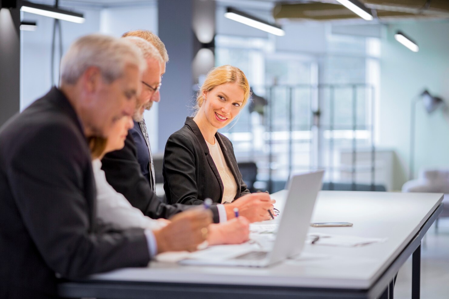 In business attire, a group of employees sits at a long desk reviewing a laptop screen in a well light office building. 